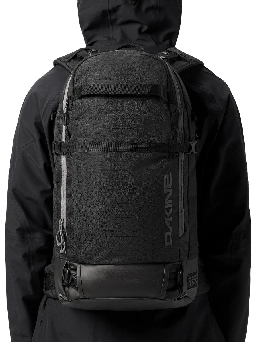 Person in a black hooded jacket wearing the Dakine Heli 2.0 Pro Pack 26L in Black, rear view showing how it sits on the back