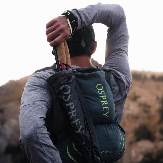 A person outdoors wearing an Osprey hydration backpack, seen from behind as they stretch their arm overhead. The backpack is dark green and black with the brand name 'OSPREY' printed vertically in light green on the side.
